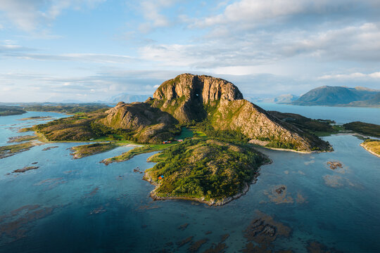 Aerial view of Torghatten mountain with its iconic natural tunnel, surrounded by the coastal landscape, islands, and clear blue waters in Bronnoy Municipality, Nordland, Norway.