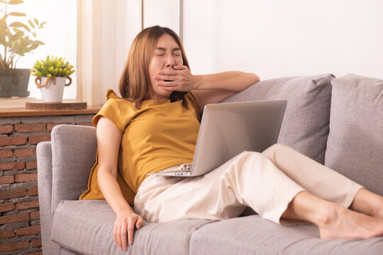 Tired woman working from home on laptop, experiencing work fatigue from prolonged screen time and sedentary work on a sofa.