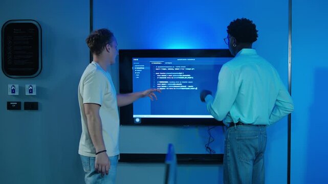 Two diverse male software developers collaborating on a coding project in a modern dark high-tech office, analyzing algorithms on a large digital screen together to solve complex software bugs.