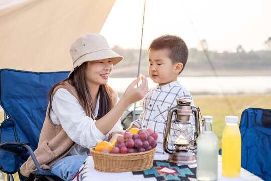 An asian mother and her young child enjoy a picnic outdoors with fruit, drinks and camping gear by a tent.