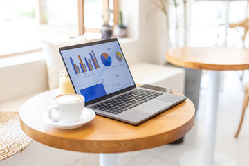 Fototapeta premium A laptop displaying business charts sits on a wooden table next to a coffee cup in a bright cafe workspace.