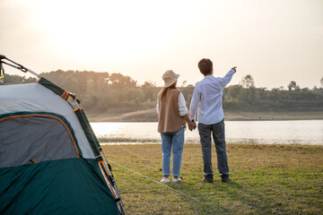 Fototapeta premium A couple holds hands while pointing at scenery by a lake at sunset, with a camping tent on the grass nearby.