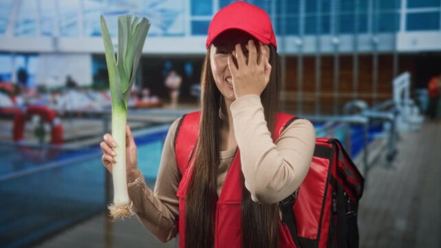 Woman courier holding a leek and covering her face with one hand in front of a glass building, wearing red cap and delivery backpack; embarrassment quirky moment.