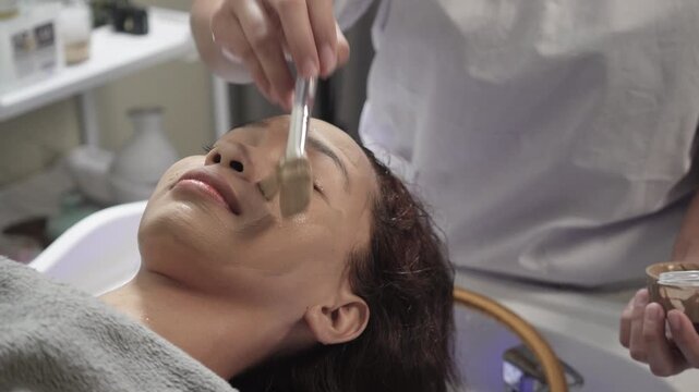 A southeast asian woman receiving a mineral mud clay facial mask application before a jade mask treatment at a Japanese head spa salon.