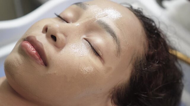 Woman receiving mineral mud clay facial mask application before professional jade mask treatment at beauty salon for Southeast Asian woman in her early 20s