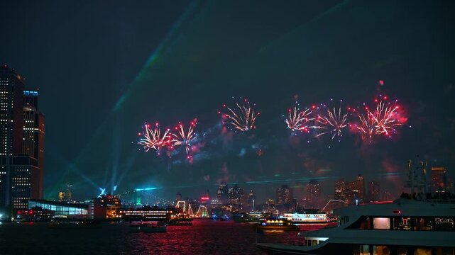 Red fireworks exploding over New York City skyline at night. Bright bursts illuminate the dark sky above illuminated passenger boats on the river during Independence Day.