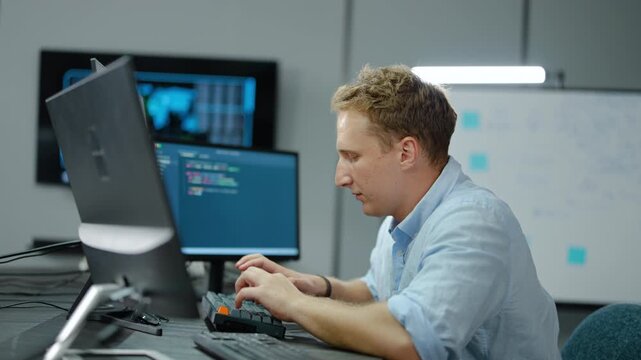 Focused IT professional working at desk typing on computer keyboard in modern control room with data charts on dual monitors. High-tech corporate workspace concept for cybersecurity or finance.