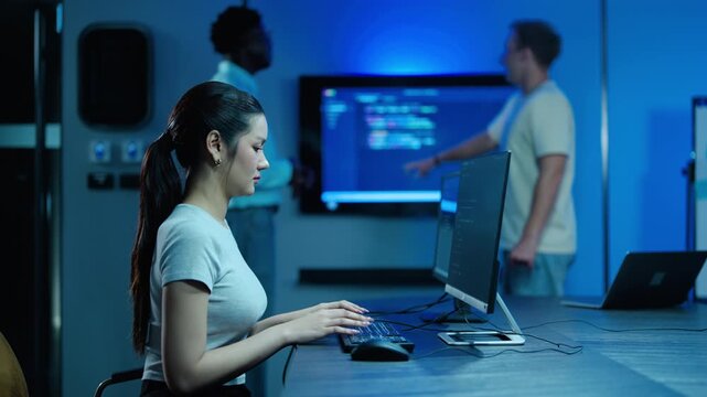 Asian female programmer typing code on a desktop computer in modern blue server room. IT team colleagues discussing software development on large screen in background. Tech business innovation.
