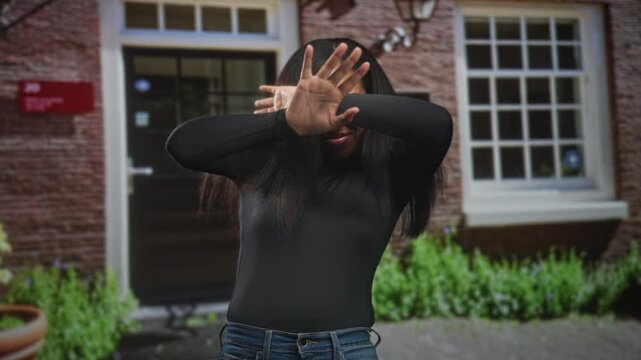 Woman covering face with hands and raised palm blocking a camera at a brick building entrance, wearing black top and jeans; privacy refusal.