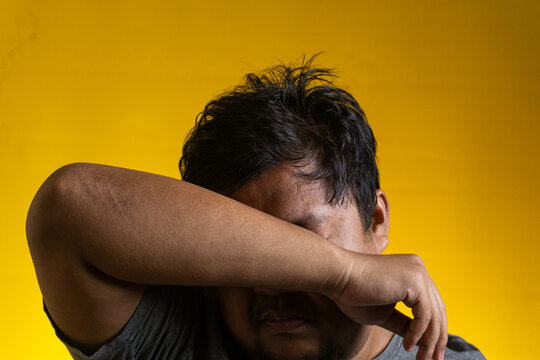 An emotional portrait of a young Asian male hiding his face by burying it into the crook of his folded arm. The subject appears overwhelmed, exhausted, or deeply saddened, positioned