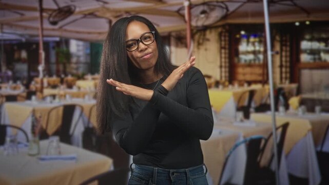 Woman wearing glasses crossing hands in an x gesture inside a restaurant building; rejection boundary assertion.