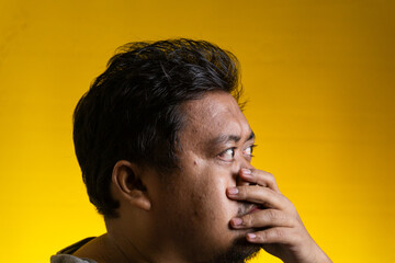 Striking profile portrait of a young Asian male covering the mouth with one hand against a bright yellow background. The individual looks intensely to the side, projecting emotions © M. Faisal Riza