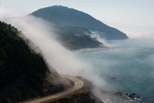 Aerial view of Highway 101 winding along the foggy Pacific coastline with lush green cliffs and ocean waves near Humbug Mountain, Oregon, United States.