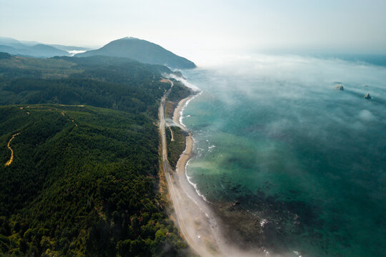 Aerial view of Highway 101 winding along the rugged coastline with dense evergreen forests, misty ocean waves, and rocky sea stacks near Humbug Mountain, Southern Oregon, United States.