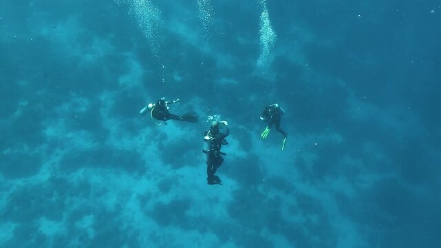Three scuba divers perform a decompression stop in open water. Two check remaining time on their dive computers while the third reels in a deployed surface marker buoy line.