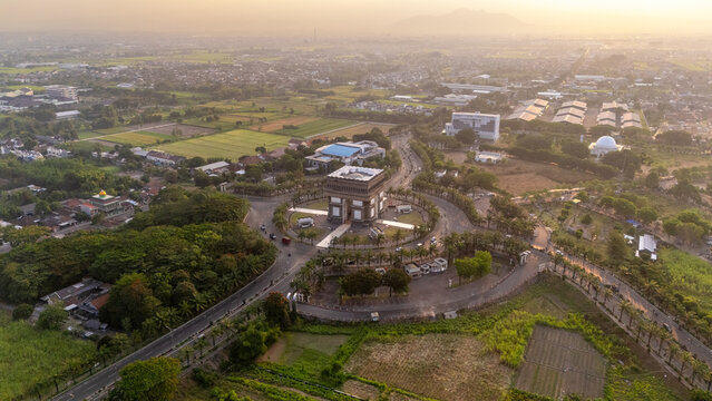 Aerial view of the Simpang Lima Gumul Monument surrounded by a roundabout, lush green fields, and palm trees during golden hour in Kediri, East Java, Indonesia.