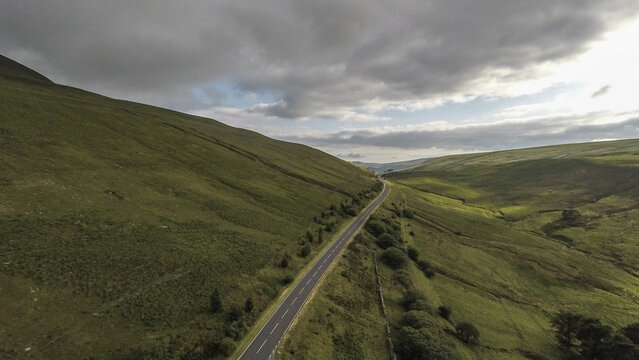 Aerial view of the Brecon Beacons with a winding road cutting through lush green hills under a cloudy sky in Brecon, Wales, United Kingdom.