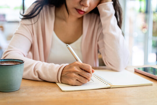 Cropped shot of a thoughtful young woman leaning her head on her hand while writing in a notebook on a wooden table