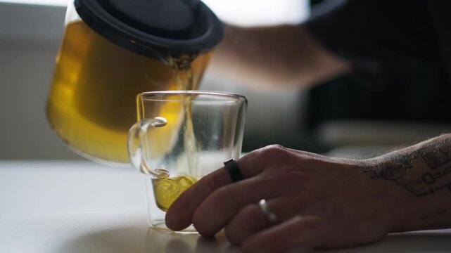 Young man with tattoos pours tea into a cup indoors.