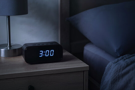 Digital alarm clock displaying three am on a nightstand. Electronic clock showing 3:00 in a dark bedroom. Insomnia, sleep disorder and early morning concept
