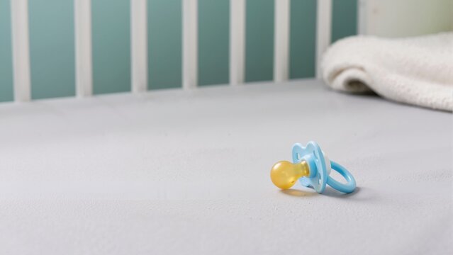 Blue baby pacifier on gray crib sheet. Dummy on mattress in nursery with wooden crib slats in background