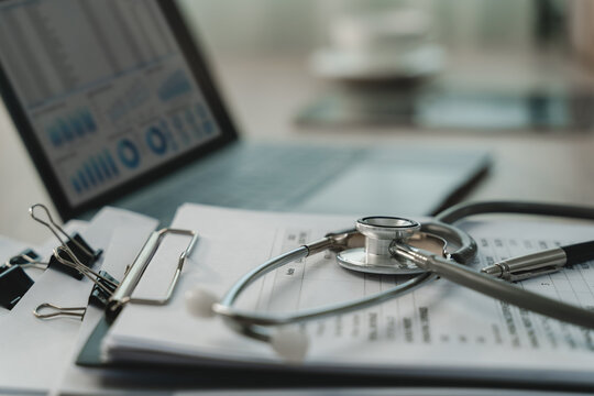 Stethoscope resting on printed medical forms beside a clipboard, pen and laptop with blurred charts, illustrating healthcare documentation and clinical record management in an office.