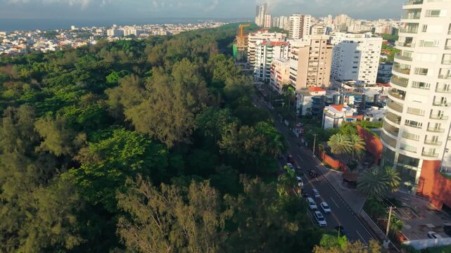 High-angle aerial drone jib shot rising over Anacaona Avenue traffic, revealing the lush Mirador Sur park and the iconic cylindrical Torre Caney against the Santo Domingo cityscape