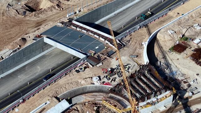 Drone video captures a highway overpass construction site with cranes, curved concrete forms, machinery, and visible workers. Bright daylight emphasizes the engineering scene.