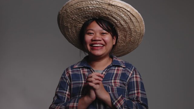 Happy young Asian female farmer wearing a straw hat and plaid shirt, clasping her hands together with a big smile.