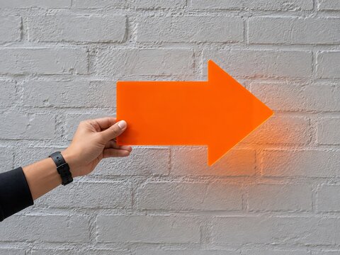 A hand holding a bright orange arrow sign pointing to the right against a textured light gray painted brick wall background surface