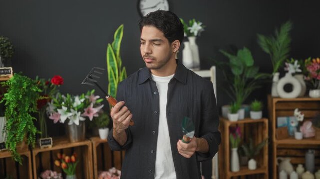 Man florist holding garden trowel and hand rake while examining tools in flower studio; quiet focus.