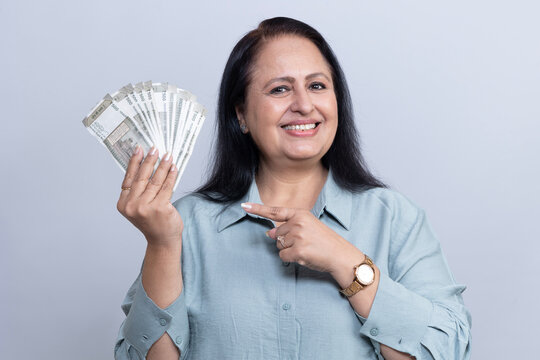 Smiling senior Indian woman holding Indian rupees notes and pointing toward the money in a studio portrait against gray background, representing savings, income, financial success, wealth.