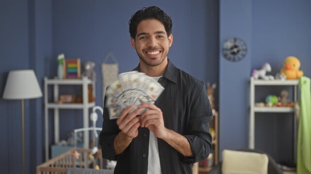 Man holding fan of dollars with both hands and smiling in a building nursery, crib and toys visible behind him; financial joy.