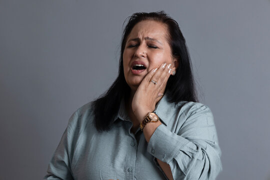 Mature Indian woman holding her cheek while experiencing intense tooth and jaw pain, with a distressed expression in a gray studio portrait, dental problems, oral health concerns,  toothache.
