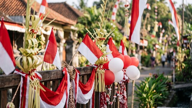Traditional Indonesian village street decorated with red and white flags, balloons, and woven coconut leaves (penjor/janur) on wooden fence, celebrating Indonesia Independence Day (17 August). 