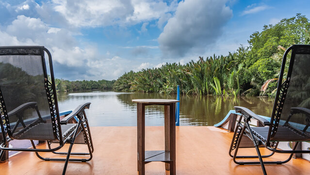 The traditional Indonesian boat klotok floats on the river in the jungle. There are two deck chairs and a table on the deck. There is a riverbed ahead, thickets of nypa fruticans palms along the banks