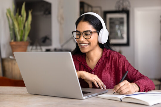 Smiling Indian student woman wearing wireless headphones studying online sits at table at home using laptop and notebook. E-learning process, remote education, professional training, online courses
