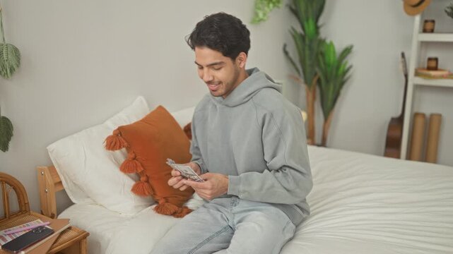 Man counting dollars with hands on bed in bedroom, seated and smiling while fanning banknotes; savings contentment.