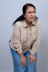 Obraz premium Mature Indian woman bending forward and holding her knee while experiencing intense leg and joint pain, showing physical discomfort and difficulty with mobility in a studio portrait.