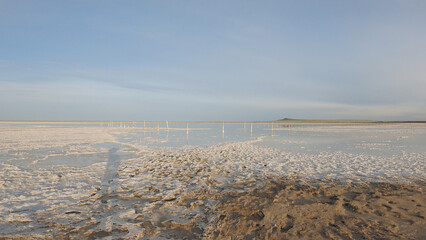 Wide view of salt flats with shallow water reflecting sky, wooden posts stretching into distance, soft golden light, textured ground, calm serene mood, ideal for travel or nature themes © VeNN