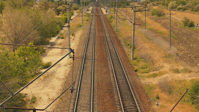 Parallel railway tracks recede into horizon under warm sunlight. Overhead power lines frame composition. Dry grass sparse green trees flank tracks. Evokes journey, travel, solitude