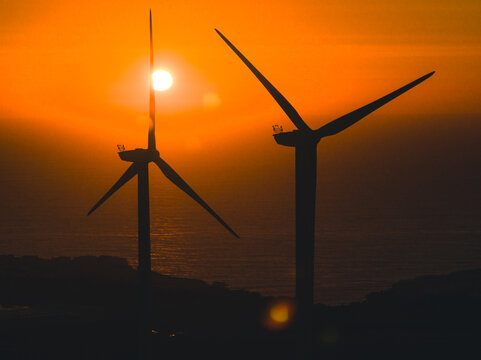 Aerial view of two wind turbines silhouetted against a vibrant orange sunset over the ocean in Santo Isidoro, Lisboa, Portugal.