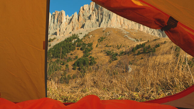 View from inside tent looking out at rugged mountain peaks under clear blue sky. Golden grass foreground, scattered evergreens on slope. Warm sunlight, natural composition, adventure mood