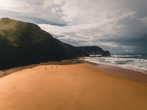 Aerial view of Praia da Cordoama with its expansive sandy beach, dramatic cliffs, and ocean waves, featuring three people walking along the shore in Lagos, Faro District, Portugal.