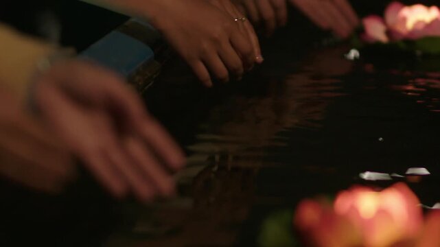 Close-up of hands placing glowing lotus lantern on dark water, traditional blessing ceremony