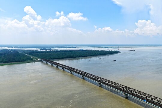 Aerial view of Jamuna Bridge spanning the wide Jamuna River with lush green riverbanks under a bright blue sky in Tangail District, Dhaka Division, Bangladesh.