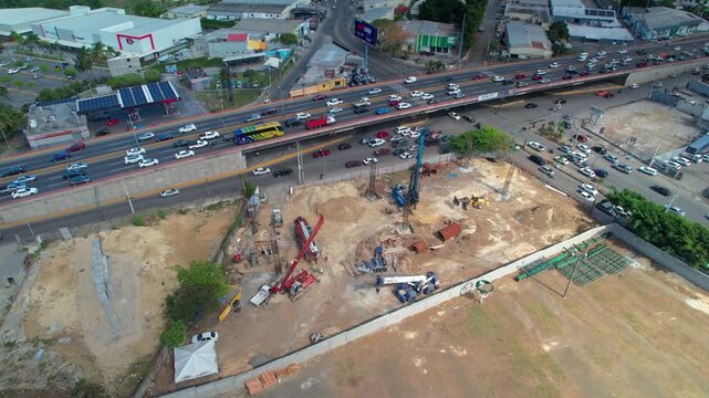 High-angle aerial drone shot pulling back and orbiting left over a massive metro infrastructure development site featuring a hydraulic piling rig and heavy highway traffic