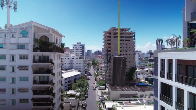 High-angle aerial drone shot ascending over an urban street canyon, showing modern residential buildings and a skyscraper under construction with a crane hoisting materials in daylight