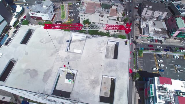 High-angle aerial drone top-down shot pulling back and rising over a tower crane hoisting materials onto a concrete skyscraper rooftop under construction in Naco