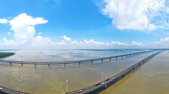 Aerial view of Jamuna Bridge spanning the wide Jamuna River with its steel truss structure and road lanes under a bright blue sky in Tangail District, Dhaka Division, Bangladesh.